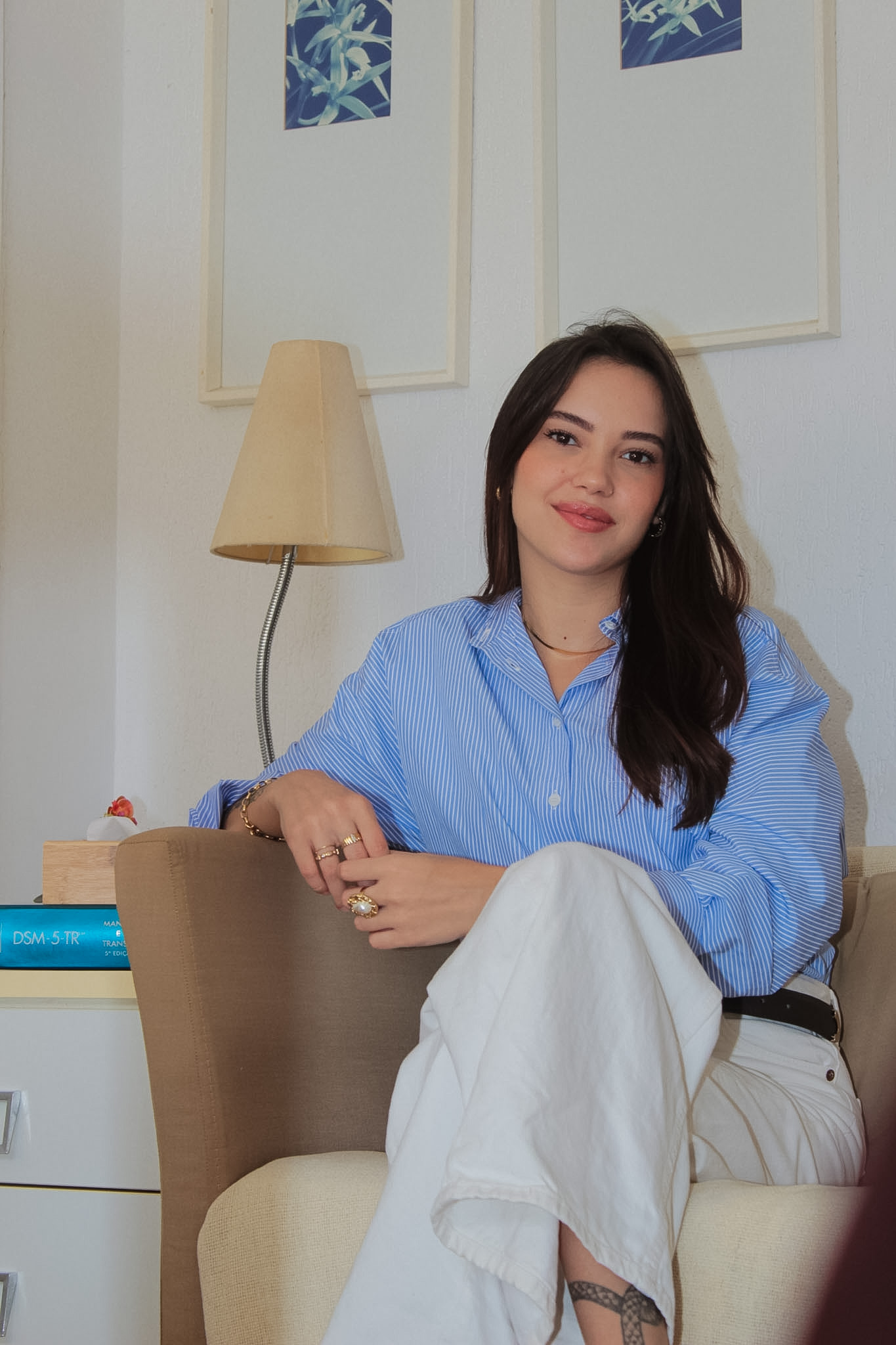 Maria Victória, psicóloga, sentada em uma poltrona no consultório, sorrindo com serenidade à luz suave da manhã